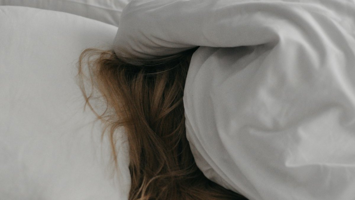 A woman is hiding under a blanket on a bed. — a Sleep Finally guide to what to do when you miss your bedtime.
