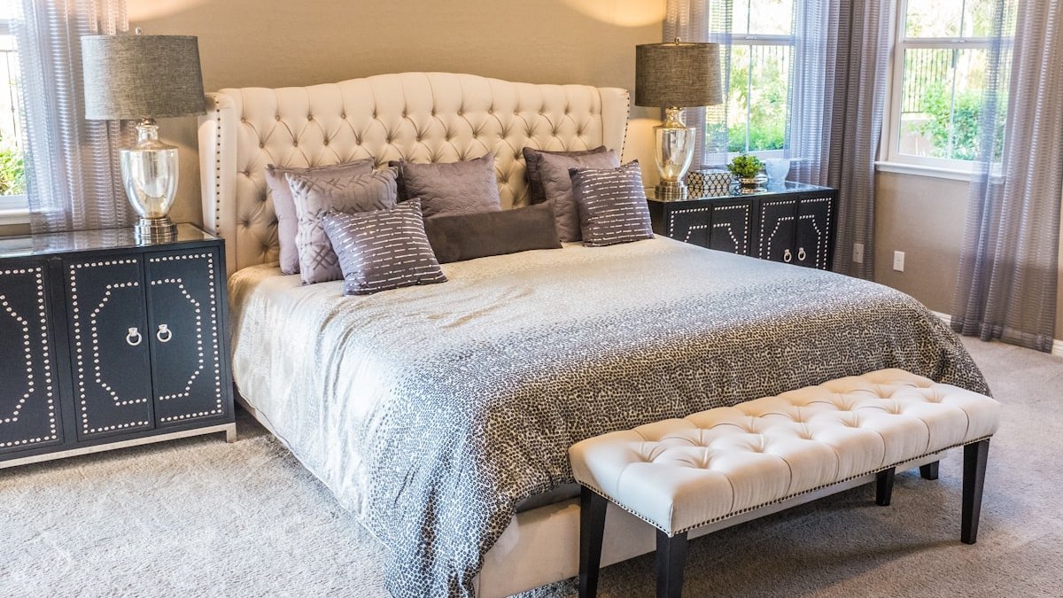 A tufted headboard bed with grey patterned bedding and a cream bench in a bedroom. — a Sleep Finally guide to Where Should the Bed Go in a Small Room? A Practical Guide.