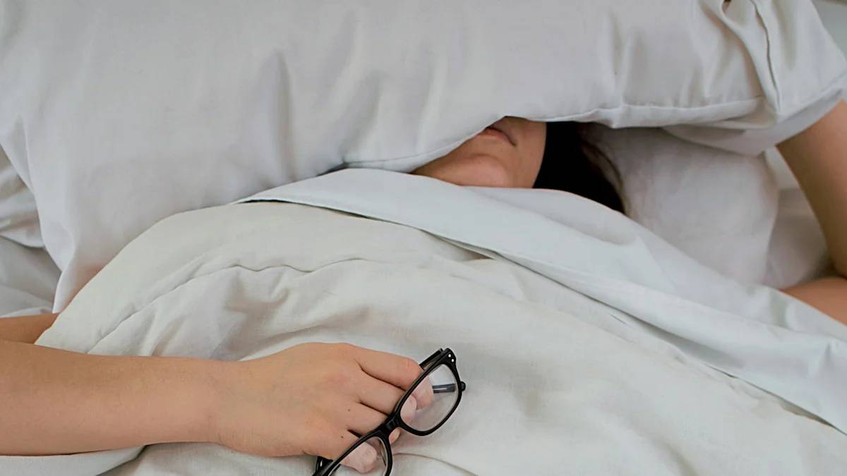 person lying on bed while covering face with pillow and holding eyeglasses — a Sleep, Finally guide to Why You Sleep 10 Hours and Still Feel Exhausted